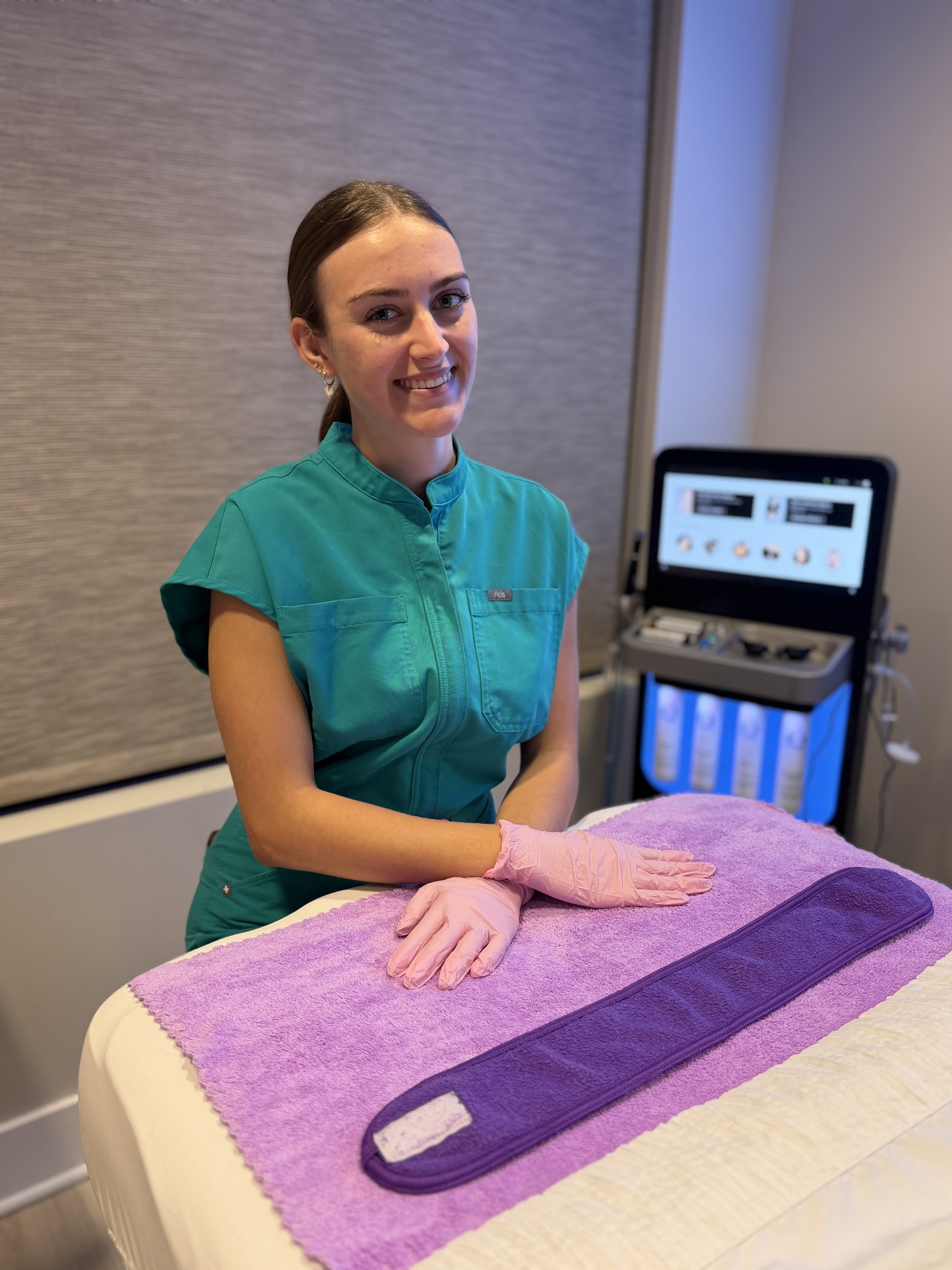 Esthetician in teal scrubs beside a HydraFacial machine